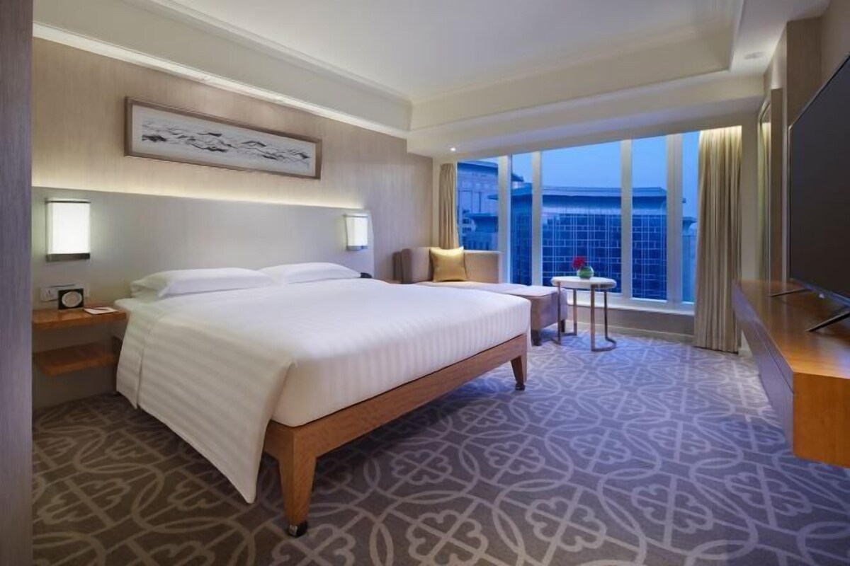 A hotel room with a large bed, a bedside table with a lamp, a small table with a vase, and a large window with a view of the city.