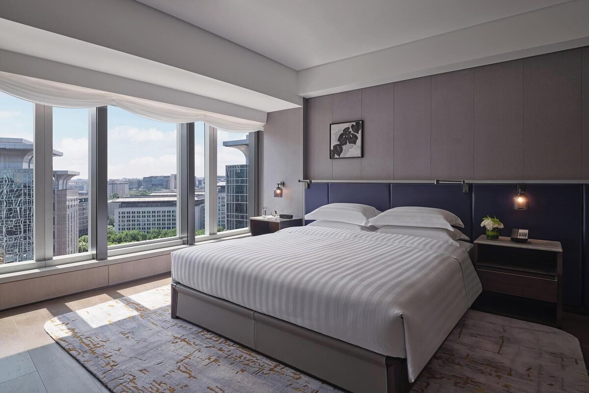 A modern bedroom with a large bed, a nightstand, a wall-mounted lamp, and a view of the cityscape through large windows.