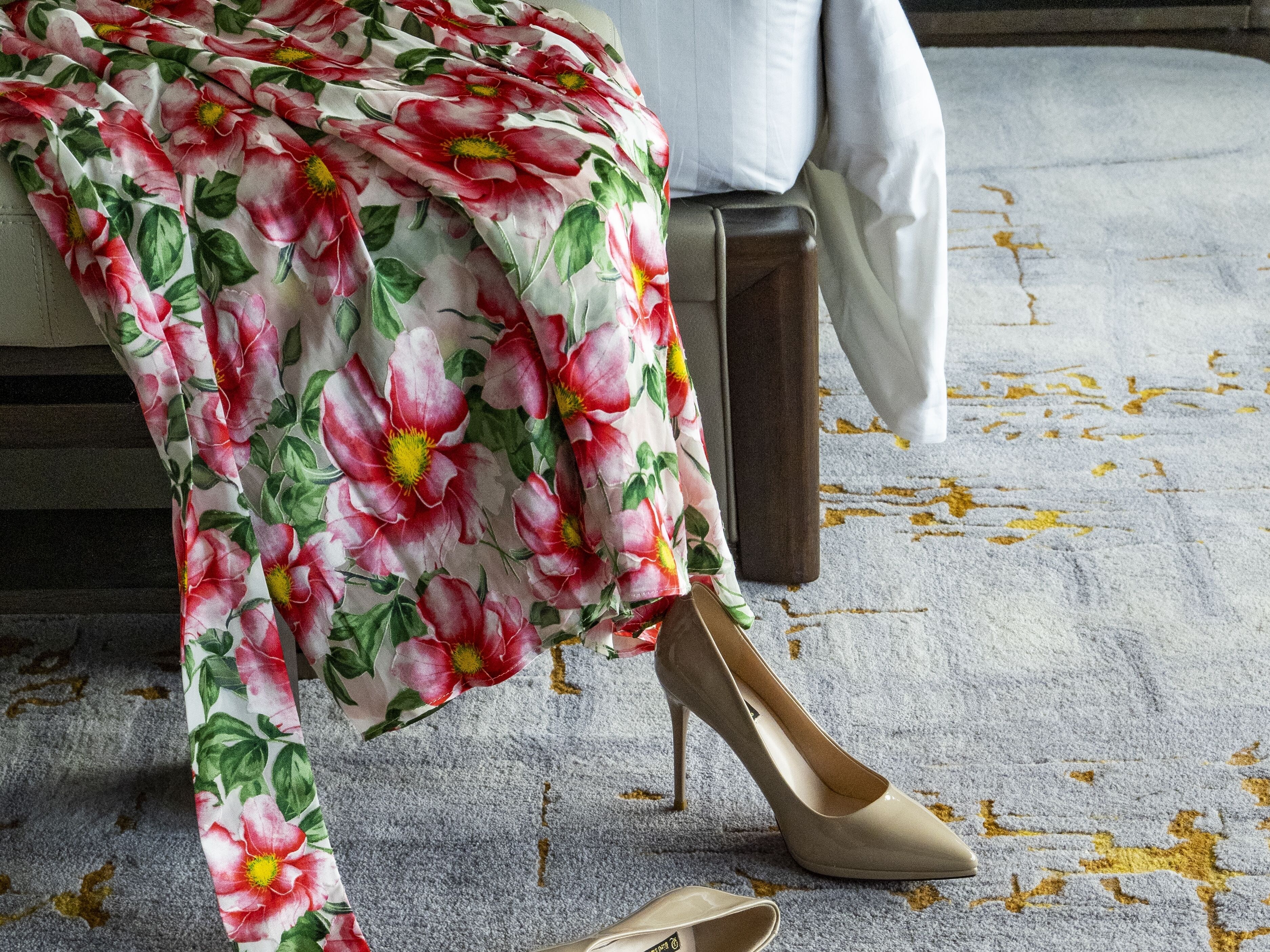 A room with a patterned carpet, a bed with a white cover, and a pair of beige high-heeled shoes on the floor.