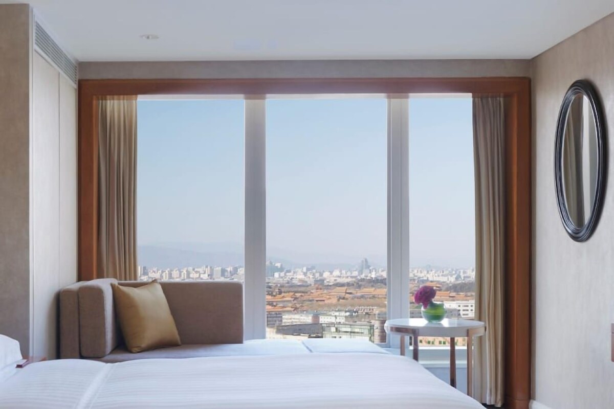 A hotel room with a large bed, a small table with a vase, and a view of a cityscape through large windows.
