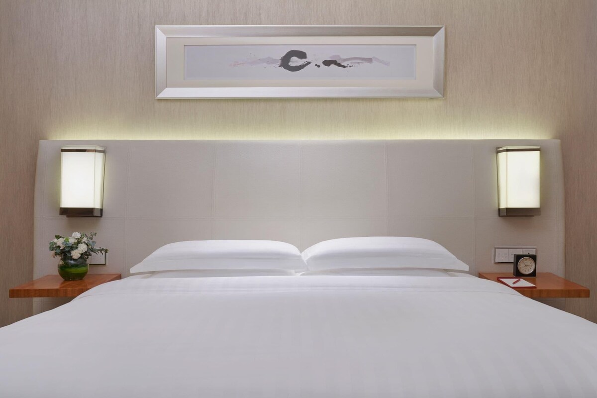 A hotel room with a neatly made bed, two bedside tables with lamps, a framed artwork above the bed, and a small clock on one of the tables.