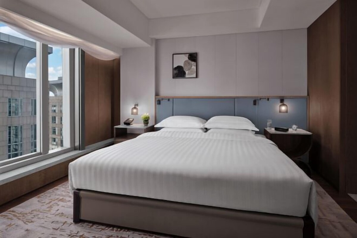 A modern hotel room with a large bed, a nightstand, a bedside table, a wall-mounted light, and a window with a view of the city.