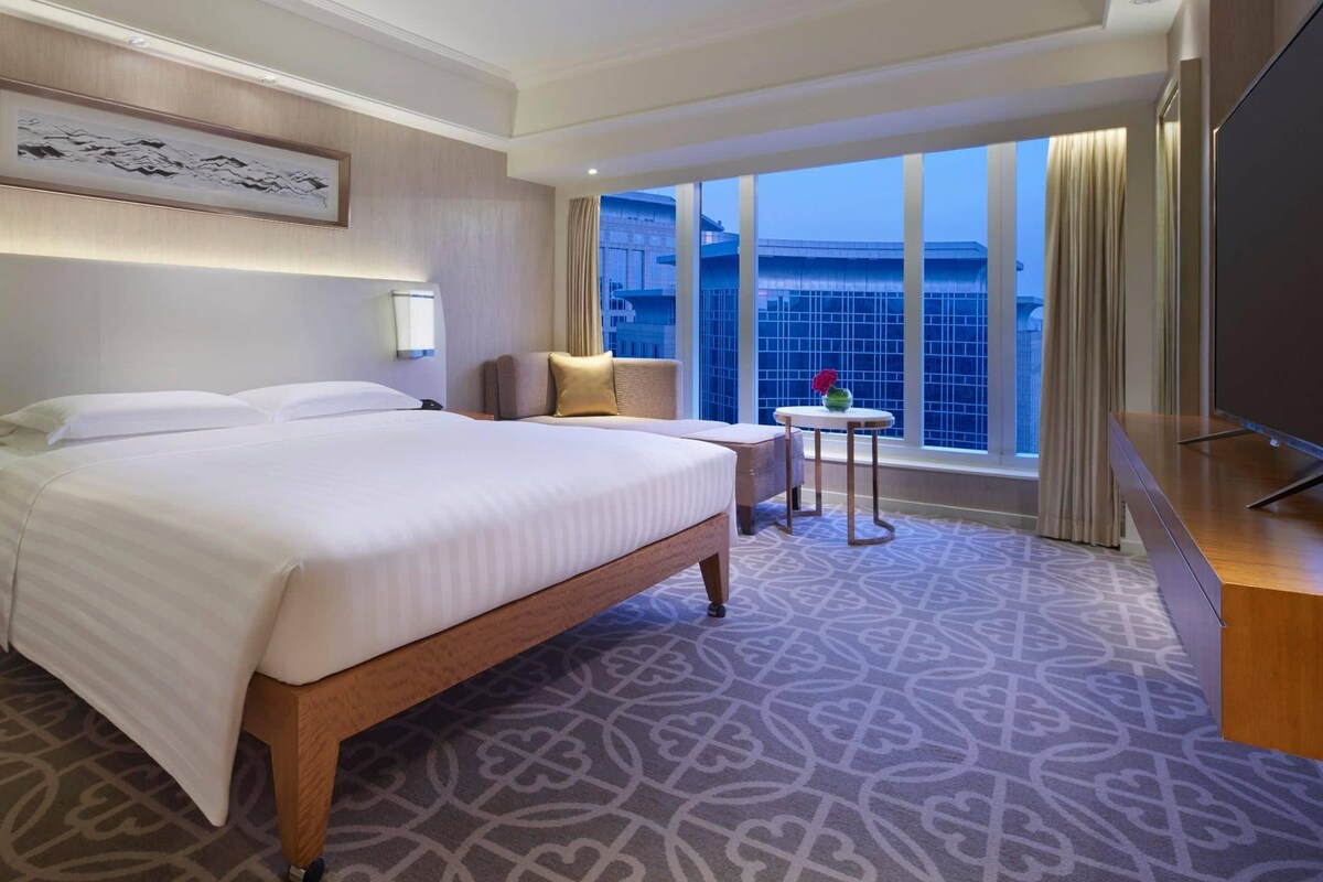 A hotel room with a large bed, bedside tables, a TV, a small table with a vase, and a window with a view of the city.