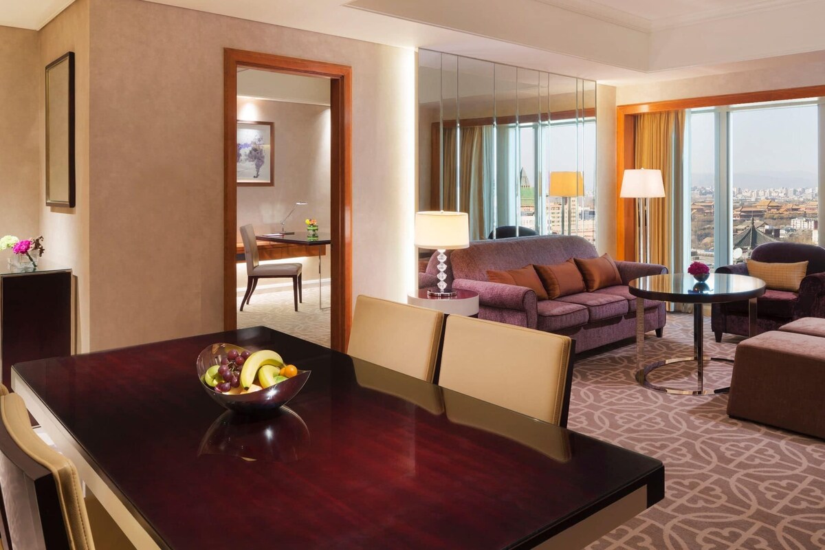 A hotel room with a dining table, a sofa, a chair, a small table with a lamp, and a large window with a city view.
