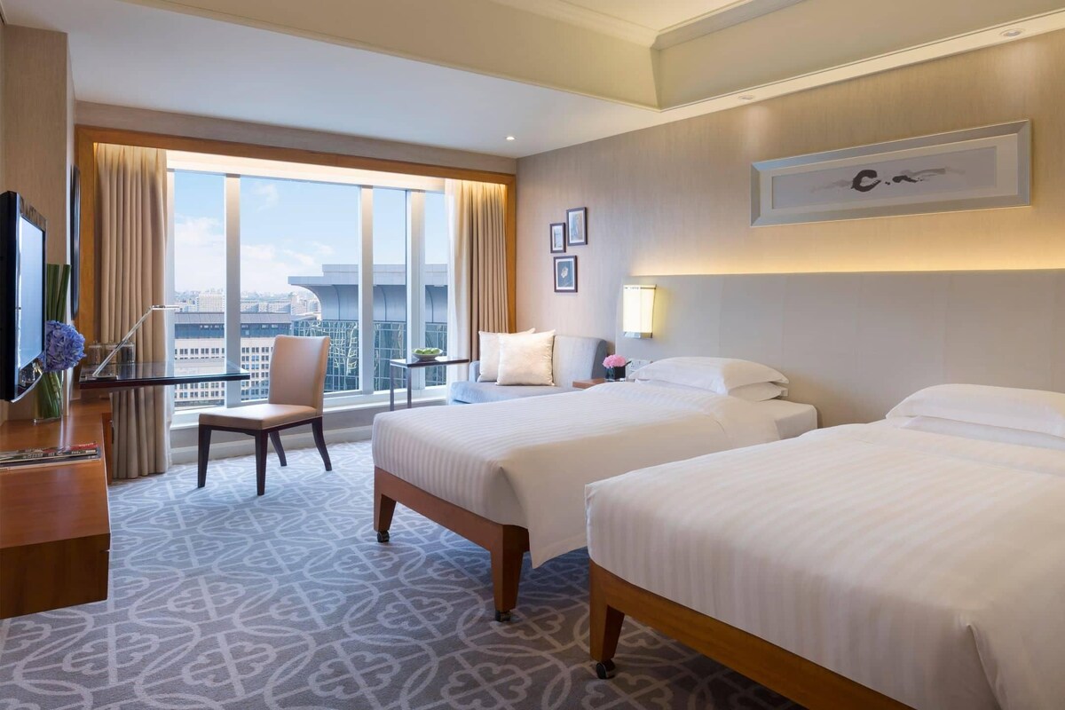 A hotel room with two beds, a desk, a chair, a TV, and a large window with a city view.