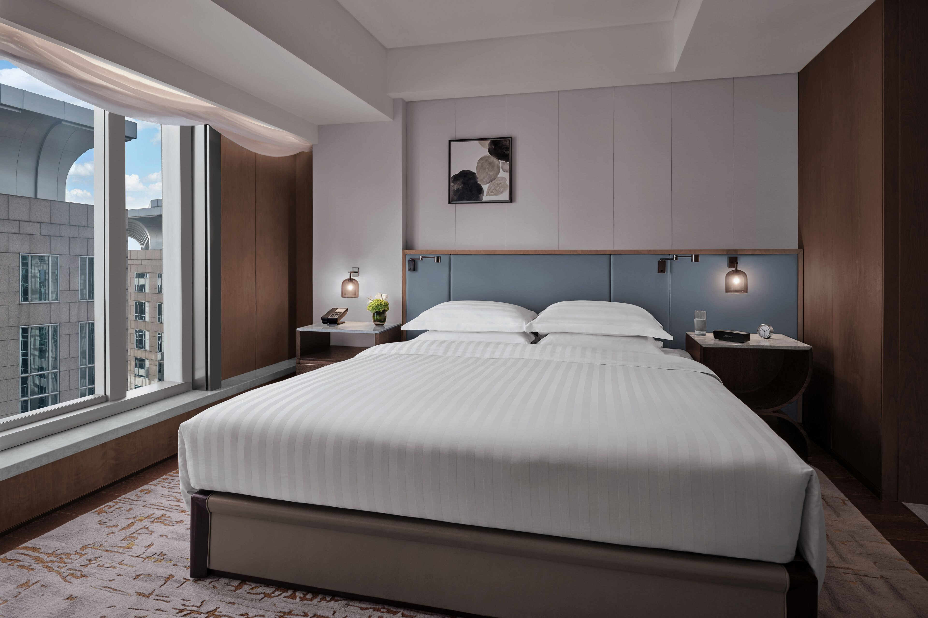 A modern hotel room with a large bed, a nightstand with a lamp, a wall-mounted artwork, and a view of the city through large windows.