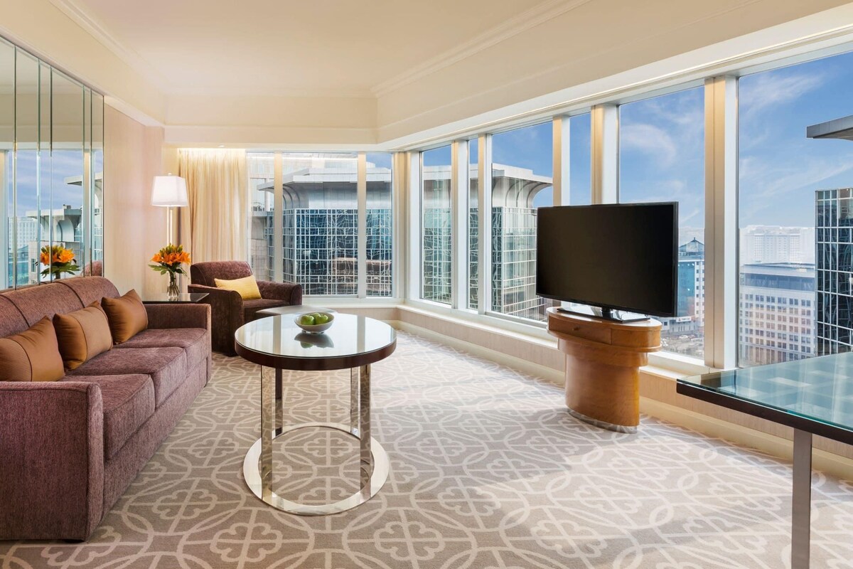 A modern hotel room with a large window, a sofa, a coffee table, and a TV.