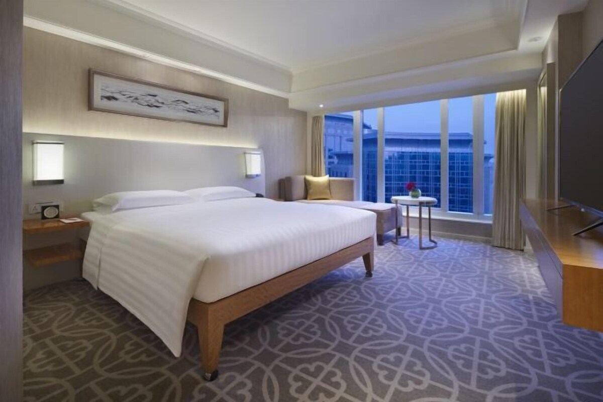 A hotel room with a large bed, a small table with a chair, a TV, and a window with a view of the city.