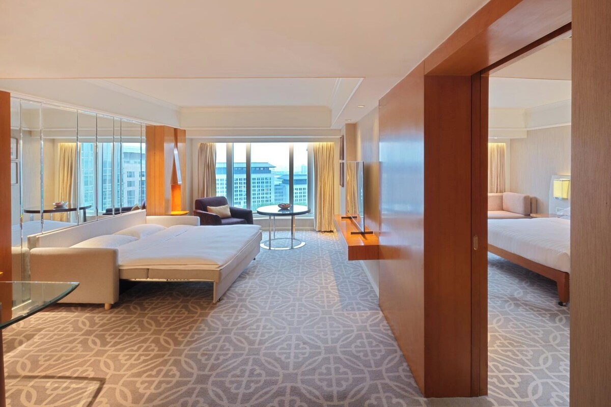 A hotel room with a large bed, a seating area with a table, a small round table, and a large window offering a city view.