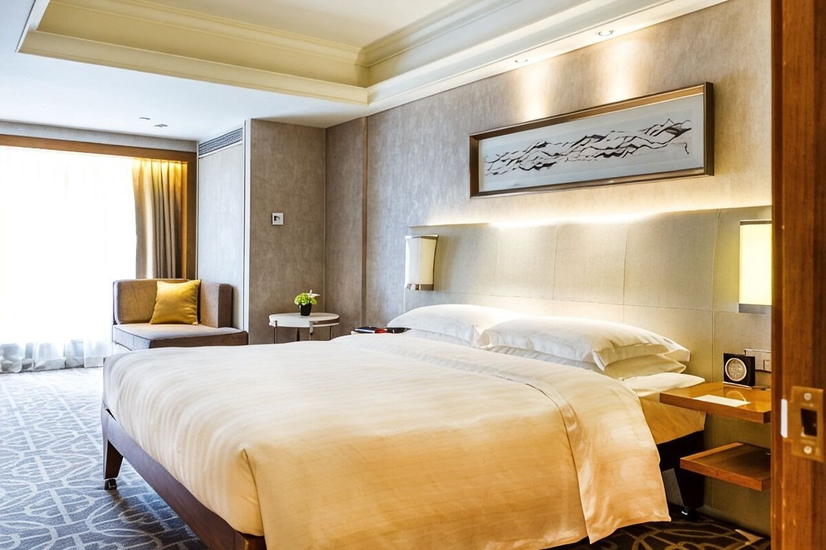 A hotel room with a large bed, a bedside table, a chair, a small table with a plant, and a wall-mounted artwork.