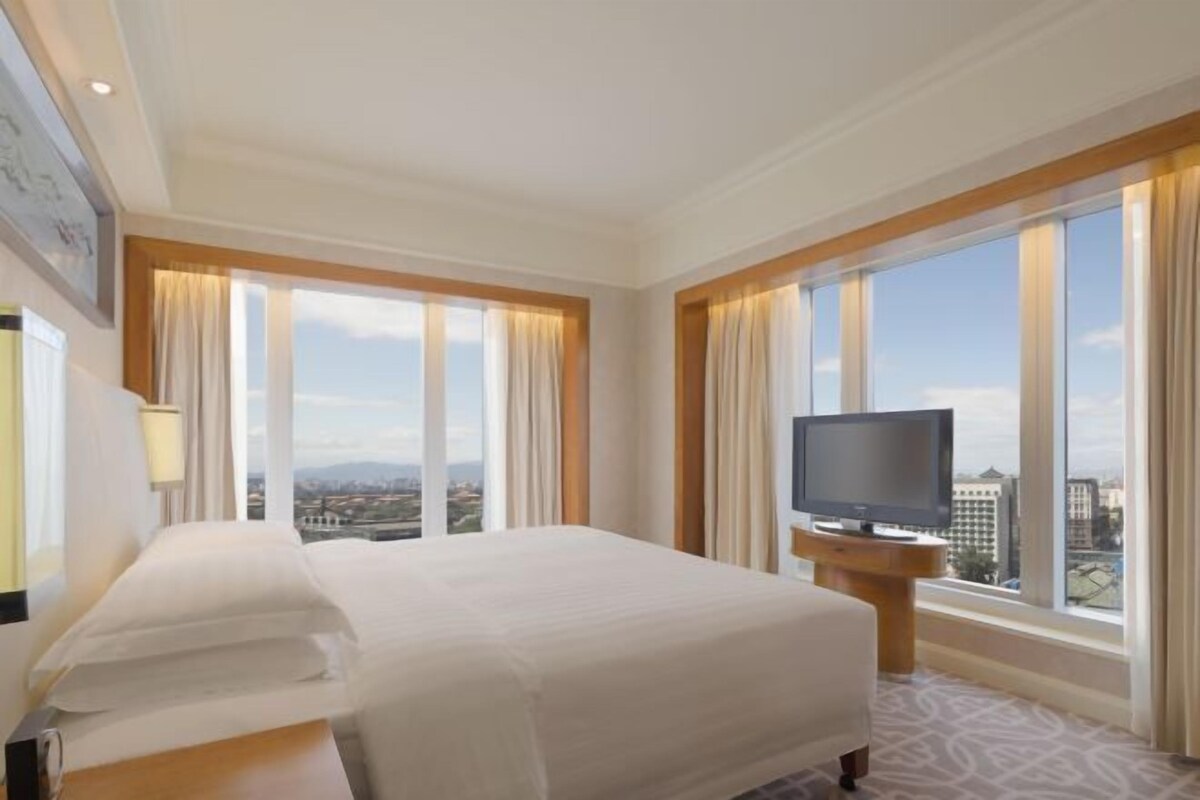 A hotel room with a large bed, a television on a wooden stand, and a view of the city through large windows.