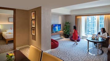 A hotel room with a bed, a TV, a dining table with chairs, a red bean bag, and a window with curtains.