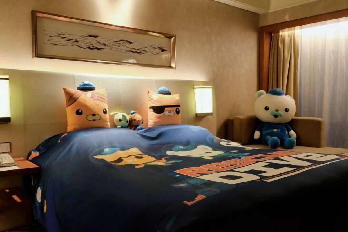 A hotel room with a bed featuring a 'Cars' themed bedspread, a large plush toy bear on the right side, and a framed artwork above the bed.