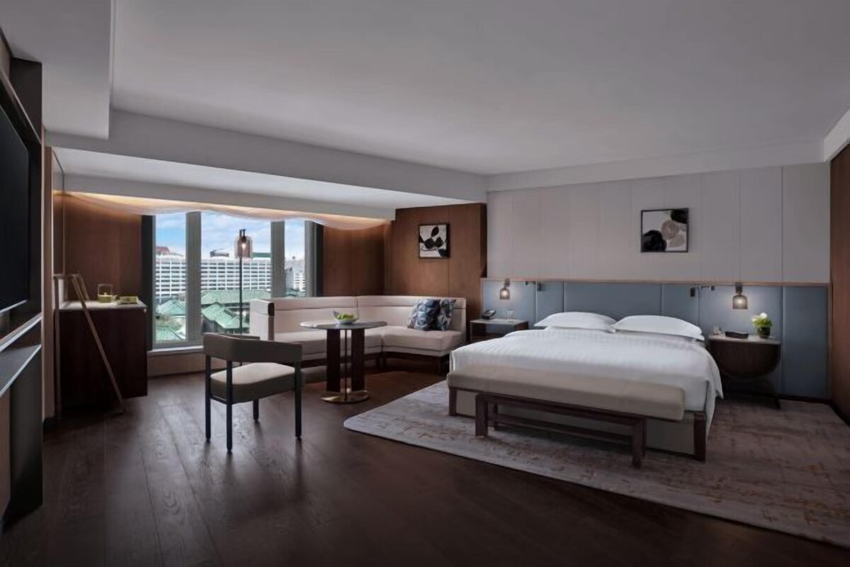A modern hotel room with a large bed, a sofa, a small table, and a chair. There is a large window with a view of buildings.