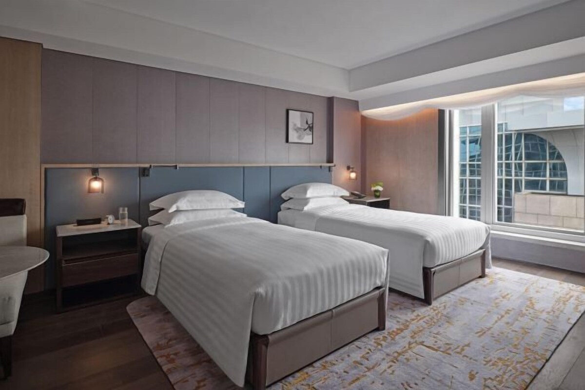 A modern hotel room with two beds, a small desk, and a large window offering a view of the city.