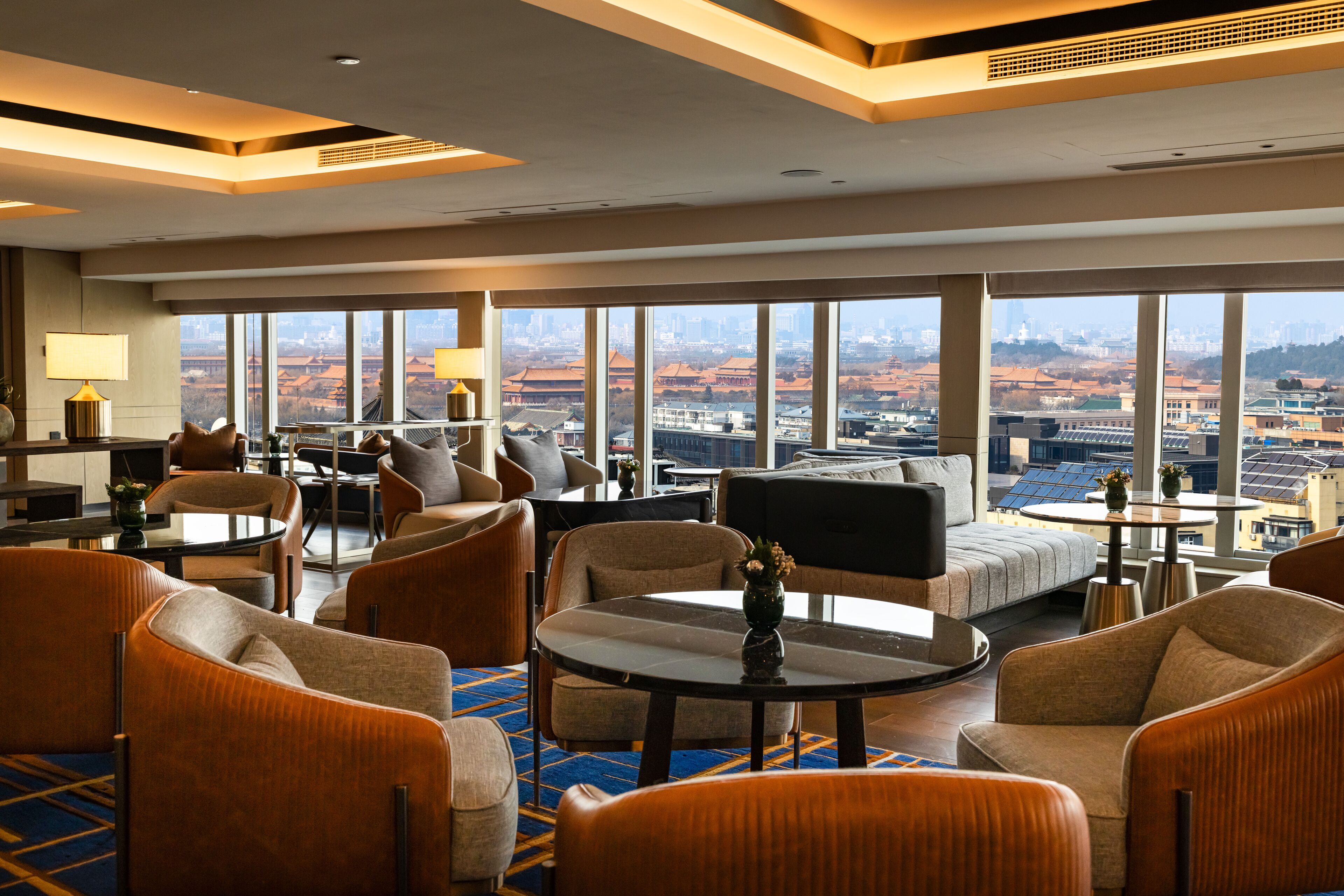 A modern hotel lobby with a large seating area, a small table, and a view of the cityscape through large windows.