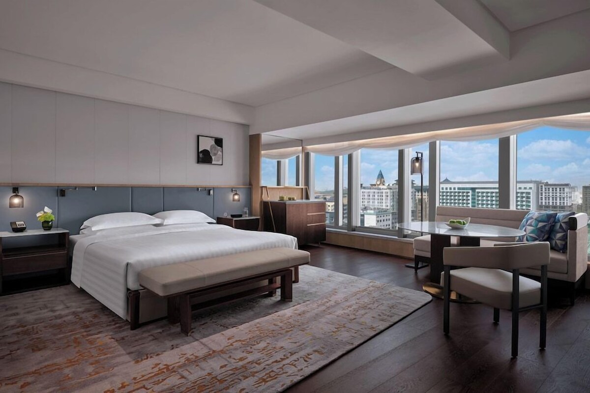 A modern hotel room with a large bed, a dining area, and a view of the cityscape.