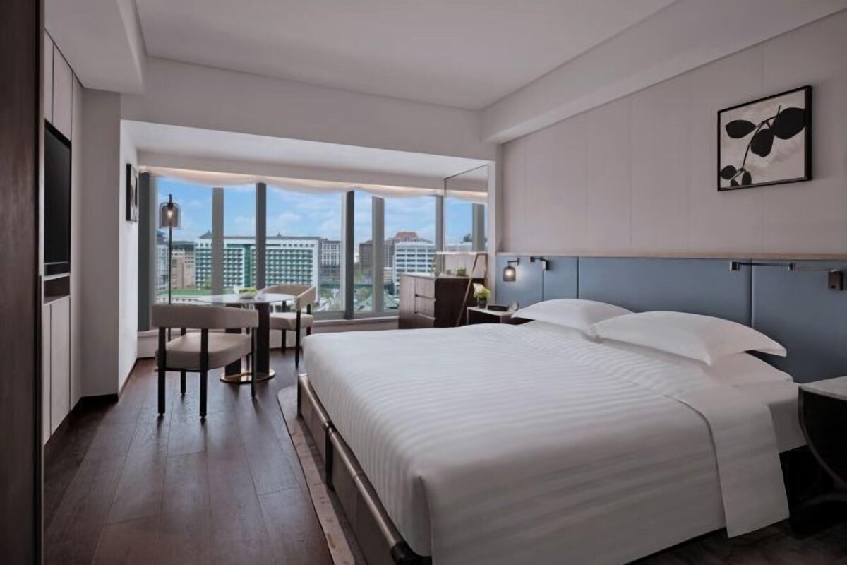 A modern hotel room with a large bed, a desk, and a chair. There is a large window offering a view of the city.