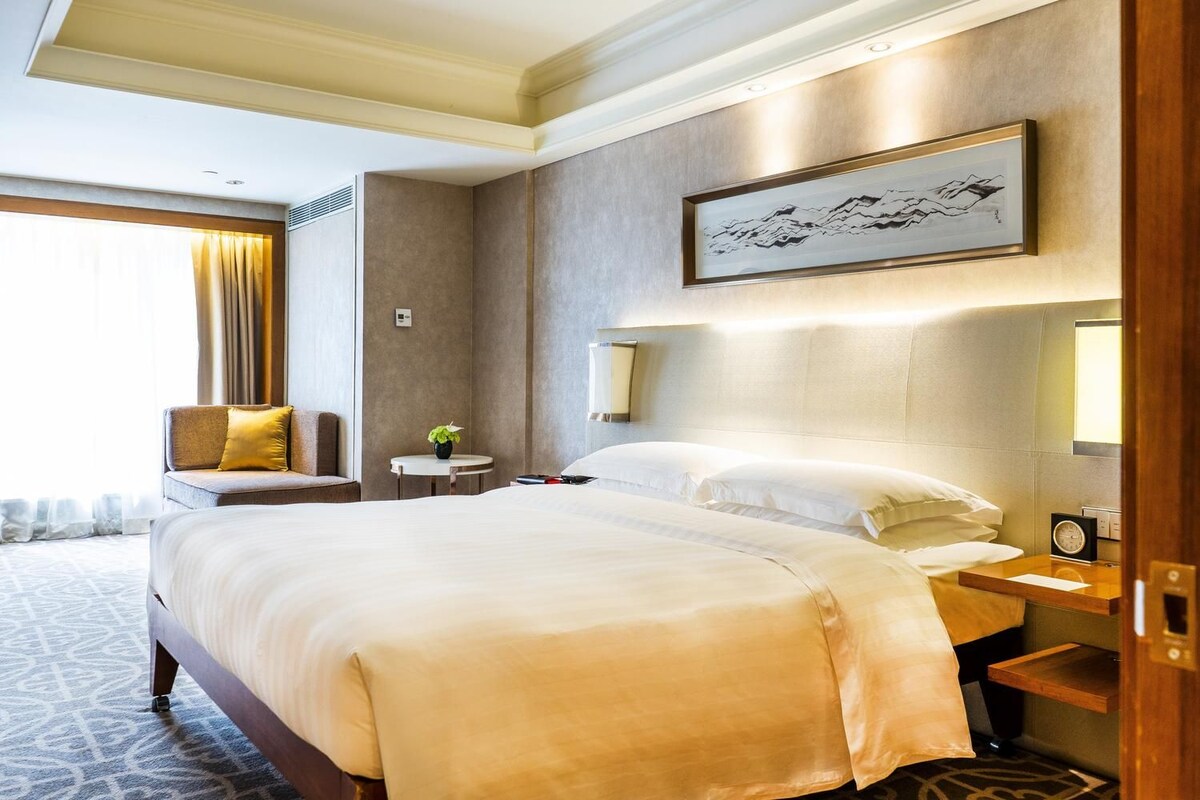 A hotel room with a large bed, a bedside table, a chair, a small table with a plant, and a wall-mounted artwork.