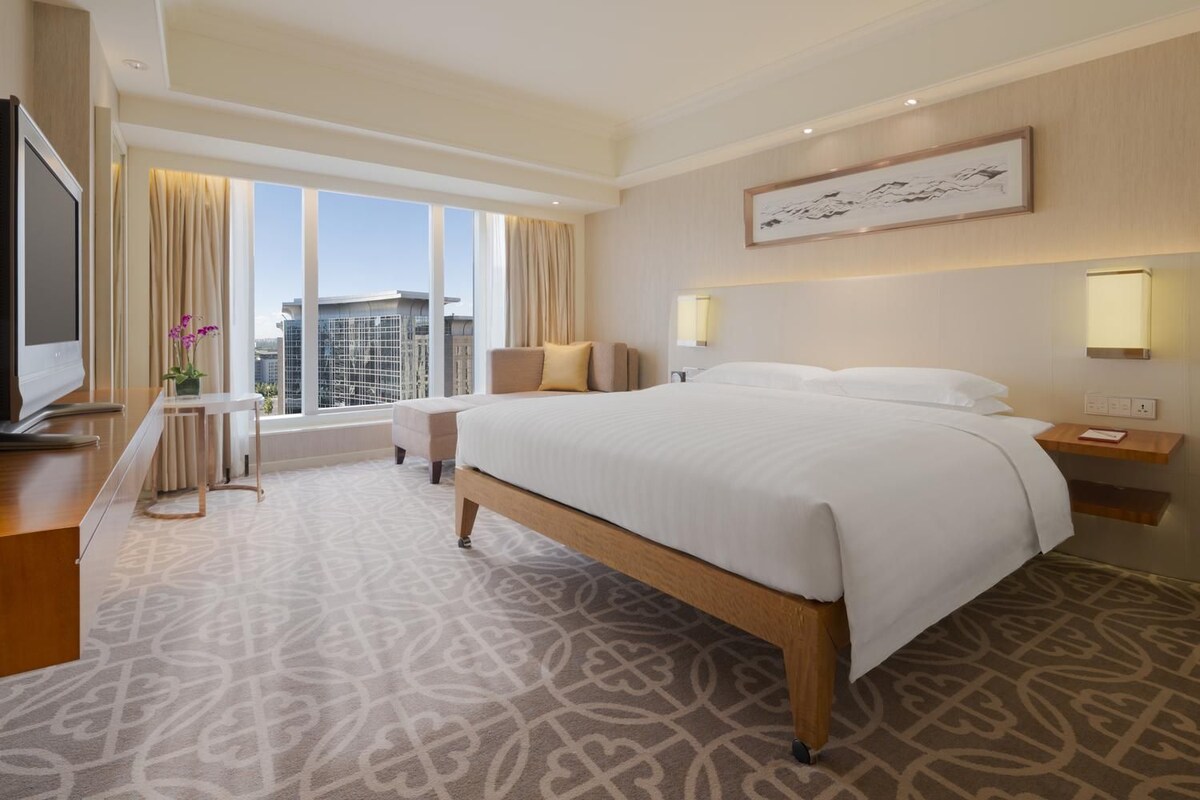 A hotel room with a large bed, a TV, a desk, and a window with curtains.