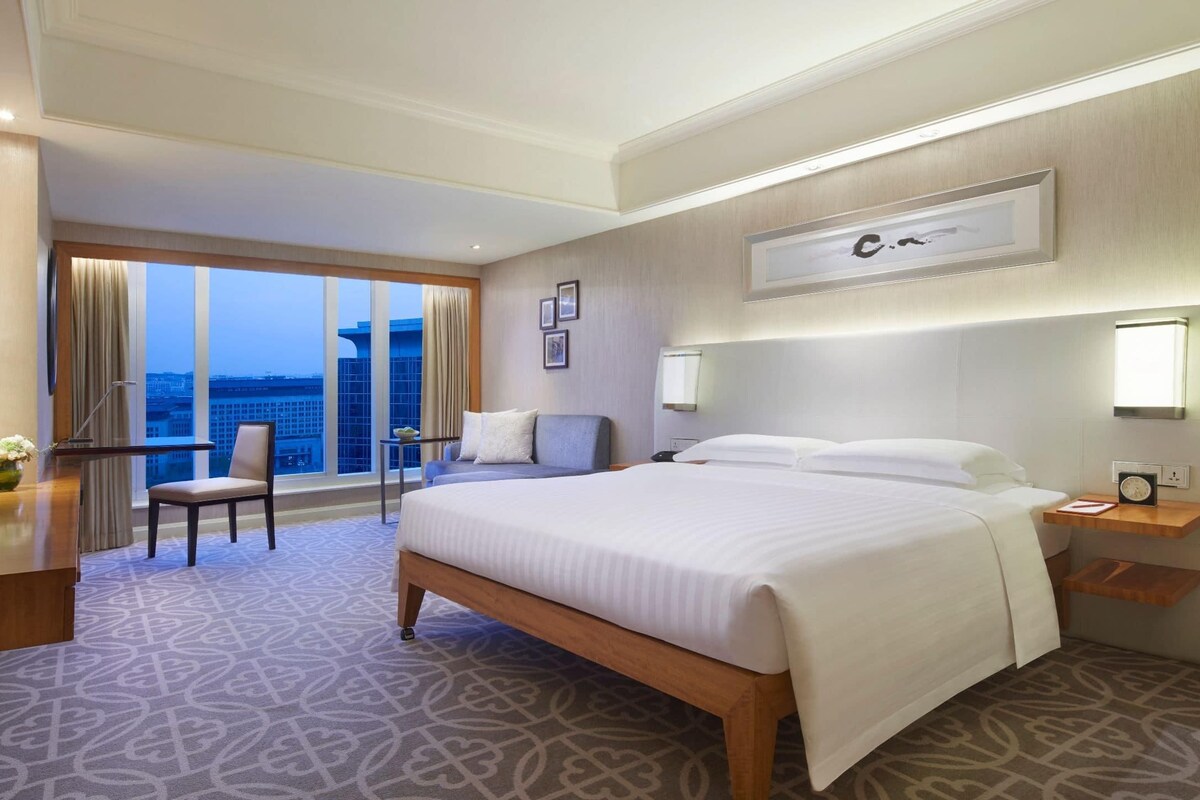 A hotel room with a large bed, a desk, a chair, a TV, and a balcony with a city view.