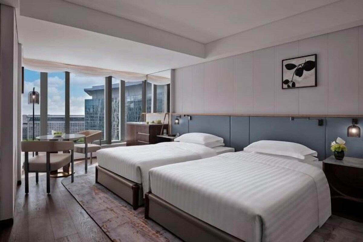 A modern hotel room with two beds, a desk, and a view of the cityscape.