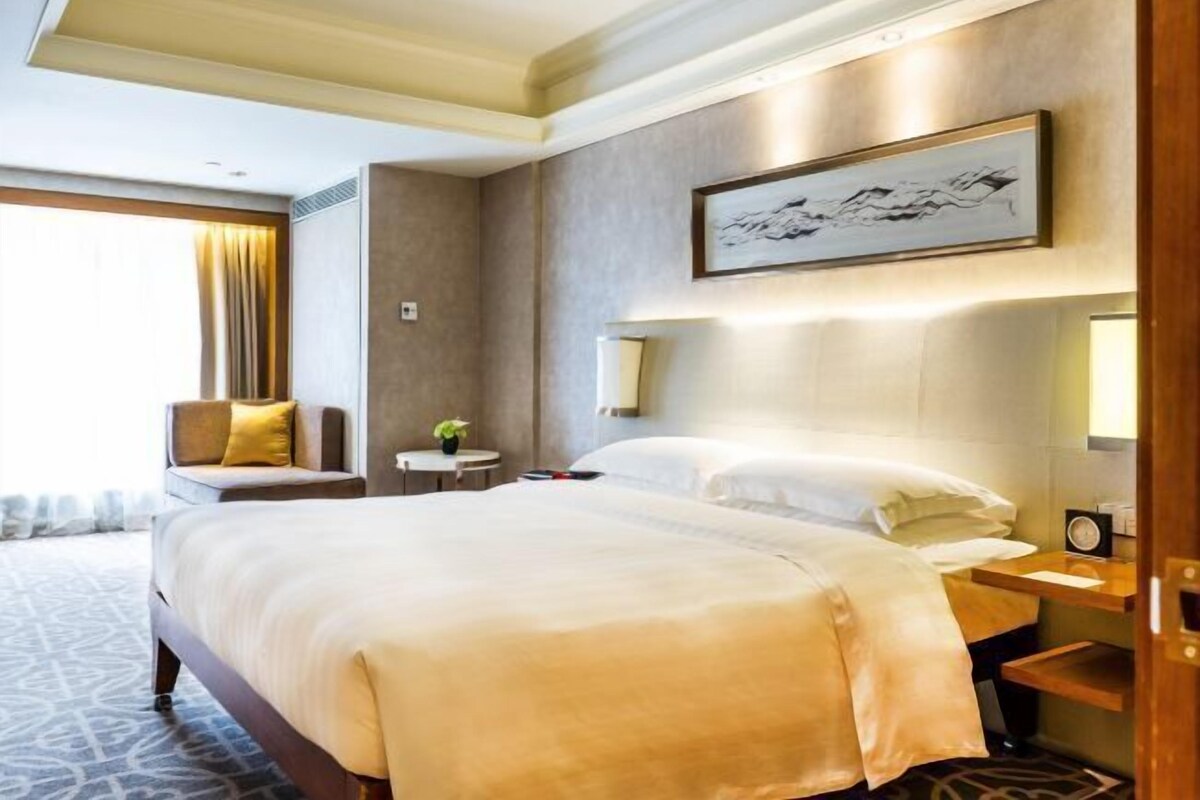 A hotel room with a large bed, a bedside table, a chair, a small table with a plant, and a painting on the wall.