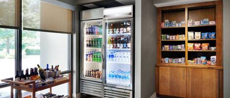 A self-service beverage station with refrigerated drinks and snacks, a wooden cabinet with packaged goods, and a window providing natural light.