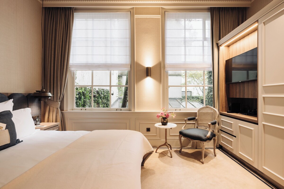 A hotel room with a large bed, a small table with a vase of flowers, a chair, and a TV mounted on the wall.