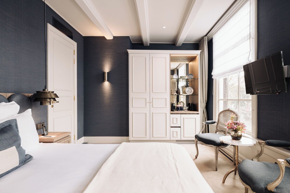 A bedroom with a large bed, a wardrobe, a vanity with a mirror, a small table with a vase, and a television on the wall.