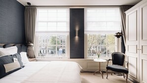 Habitación de hotel con una cama grande, una silla, una mesita y vistas a la ciudad a través de amplios ventanales.