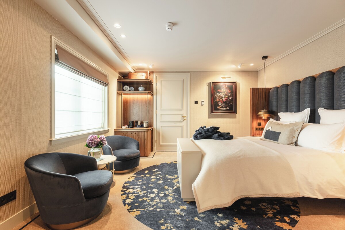 A modern hotel room with a large bed, a seating area with a chair and table, a small desk, and a decorative wall piece.