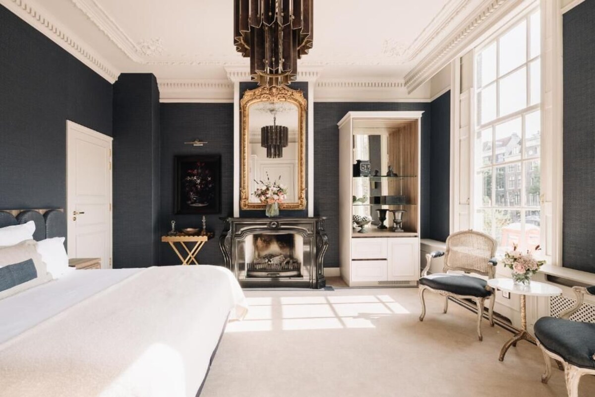A bedroom with a large bed, a fireplace, a mirror, and a view of the city through large windows.
