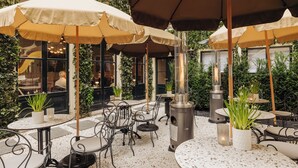 Un patio al aire libre con mesas y sillas redondas, cubierto por sombrillas beige, rodeado de follaje.
