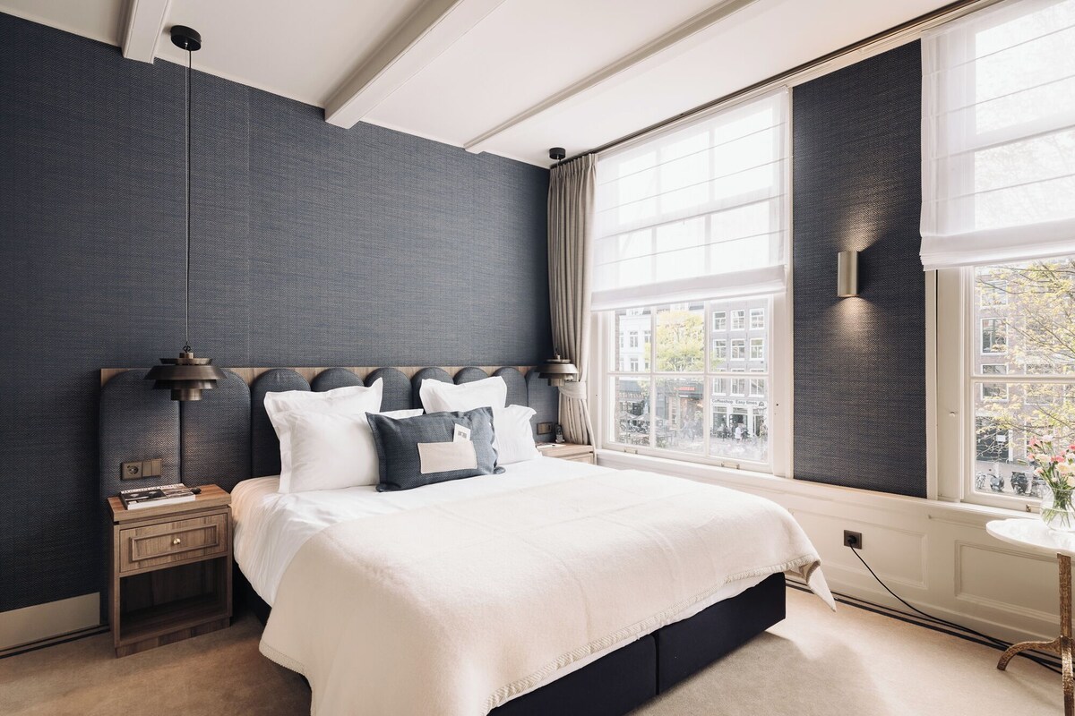 A modern bedroom with a large bed, a nightstand, a lamp, and a view of the city through a large window.