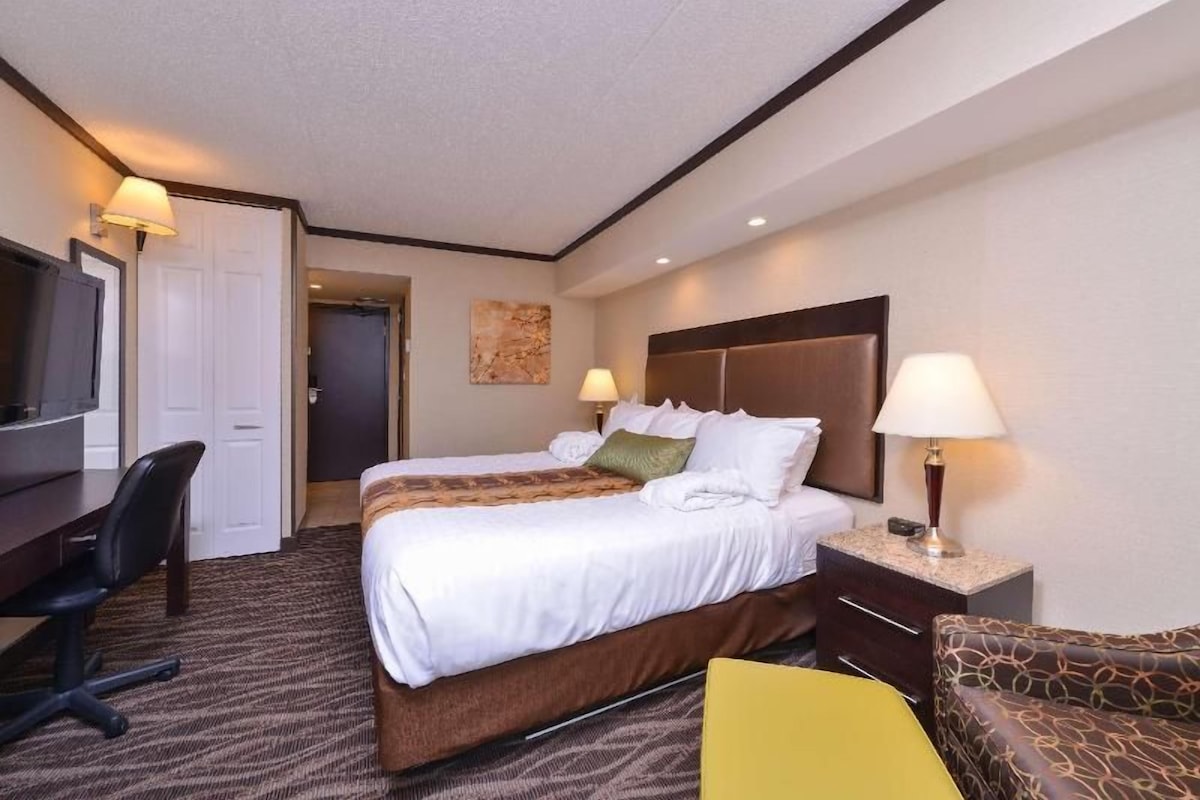 Habitación de hotel con cama, mesita de noche, lámpara, silla, escritorio, televisor y una puerta.