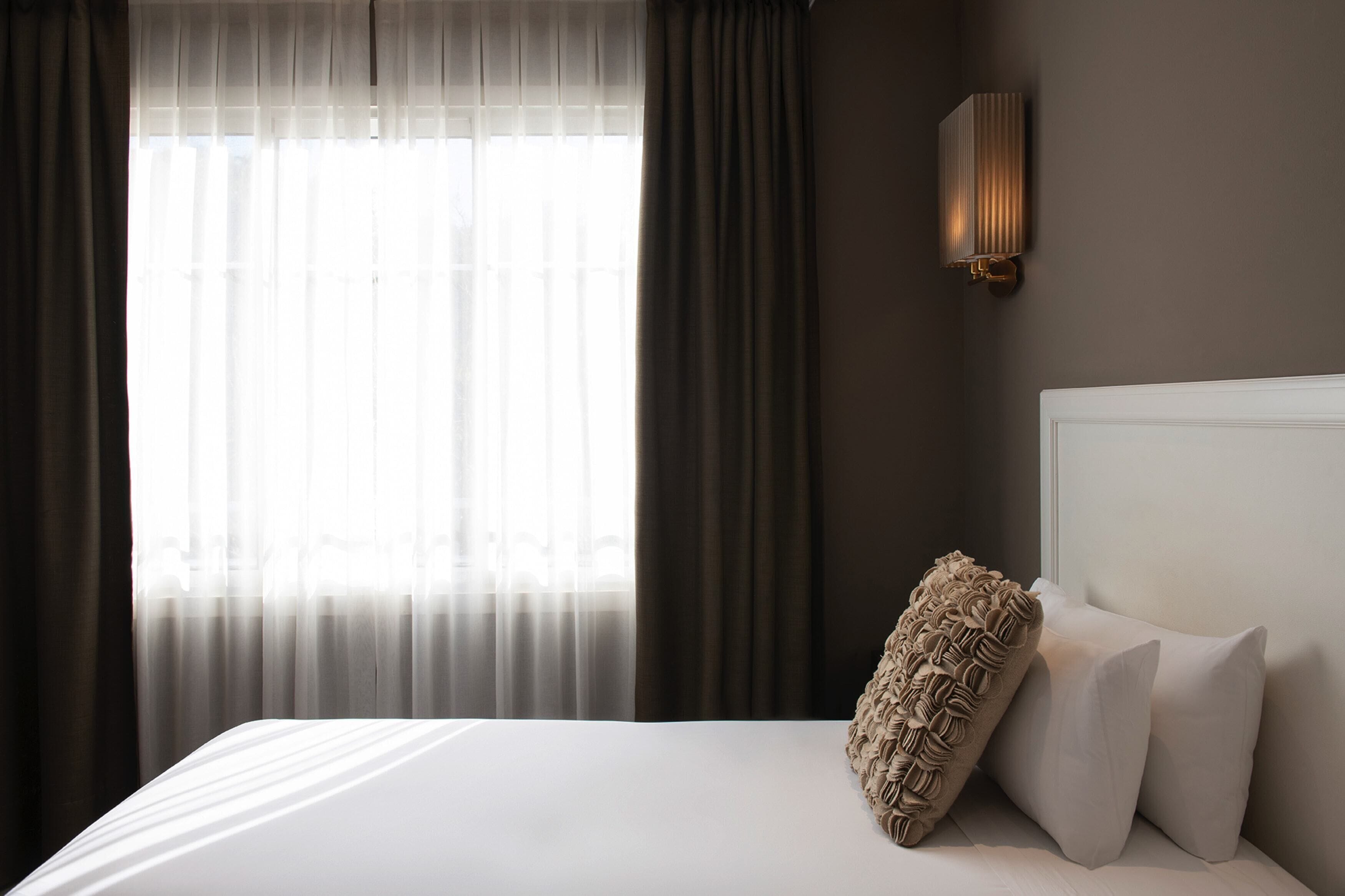 Una cama bien tendida con ropa blanca y un cojín decorativo texturizado, una lámpara de pared y cortinas traslúcidas que dejan entrar luz natural.