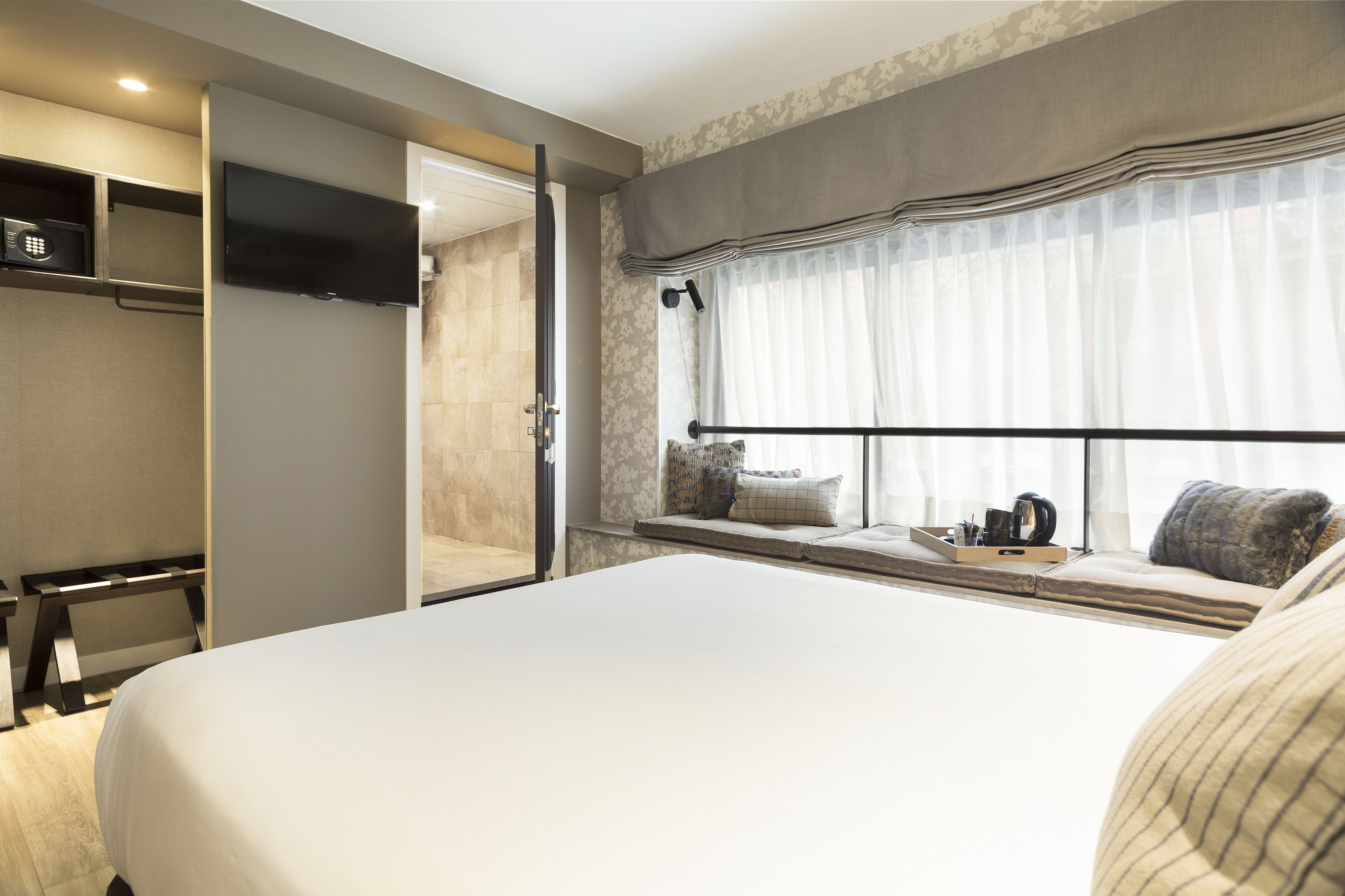 A hotel room with a large bed, a TV mounted on the wall, a window with curtains, and a bedside table with a kettle and cups.