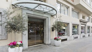 Ein Hotel mit Eingangsbereich, einer gebogenen Markise, großen Kübelpflanzen und einem sichtbaren Schaufenster mit einer Beschilderung.