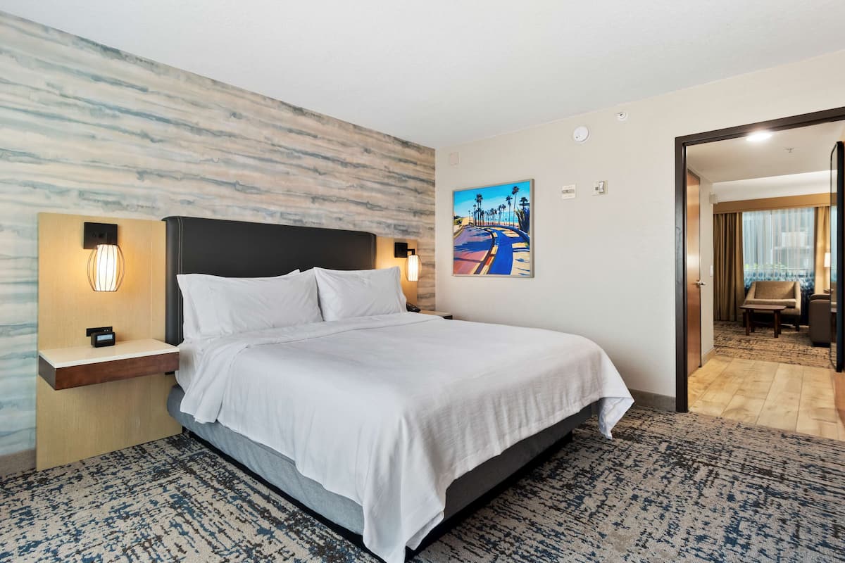 A modern hotel room with a large bed, a nightstand with a lamp, a painting on the wall, and a view into the adjacent room.