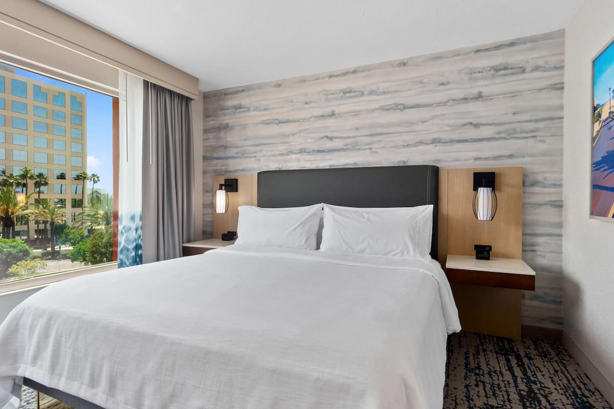 A hotel room with a large bed, a nightstand with a lamp, a view of palm trees and a building outside the window.