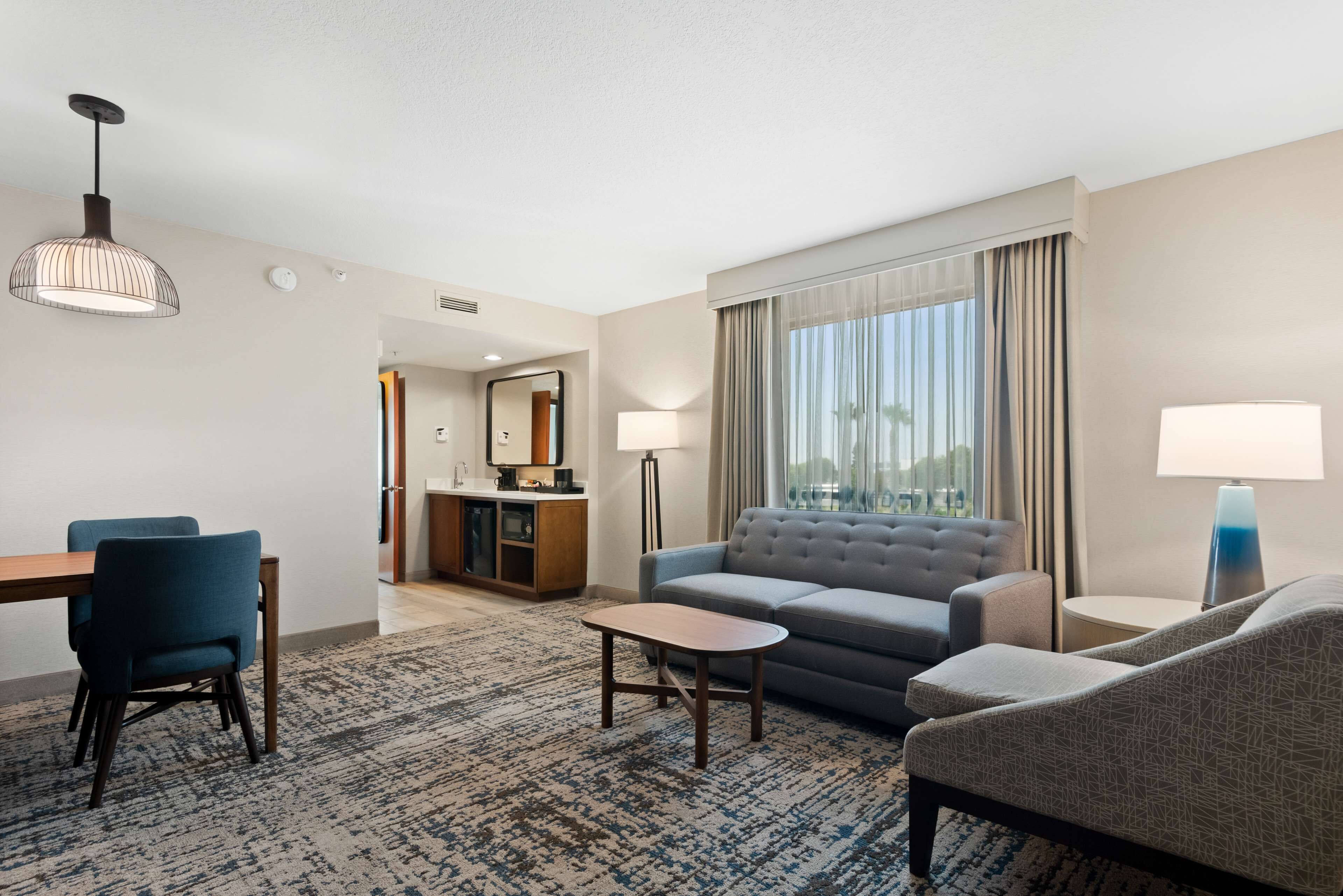 Una habitación de hotel moderna con sofá, sillones, una mesa de comedor con sillas, una pequeña mesa de centro y un ventanal con cortinas.