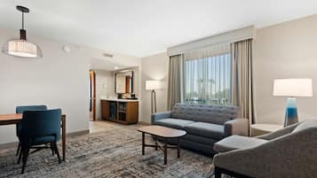 Una habitación de hotel moderna con sofá, sillones, una mesa de comedor con sillas, una pequeña mesa de centro y un ventanal con cortinas.