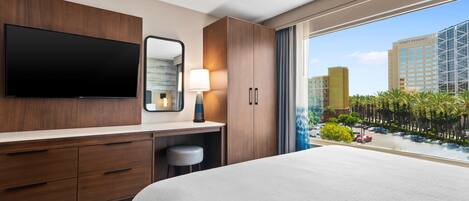 Habitación de hotel con una cama grande, un televisor de pantalla plana montado en la pared, un escritorio con lámpara y vistas a la ciudad a través de amplios ventanales.