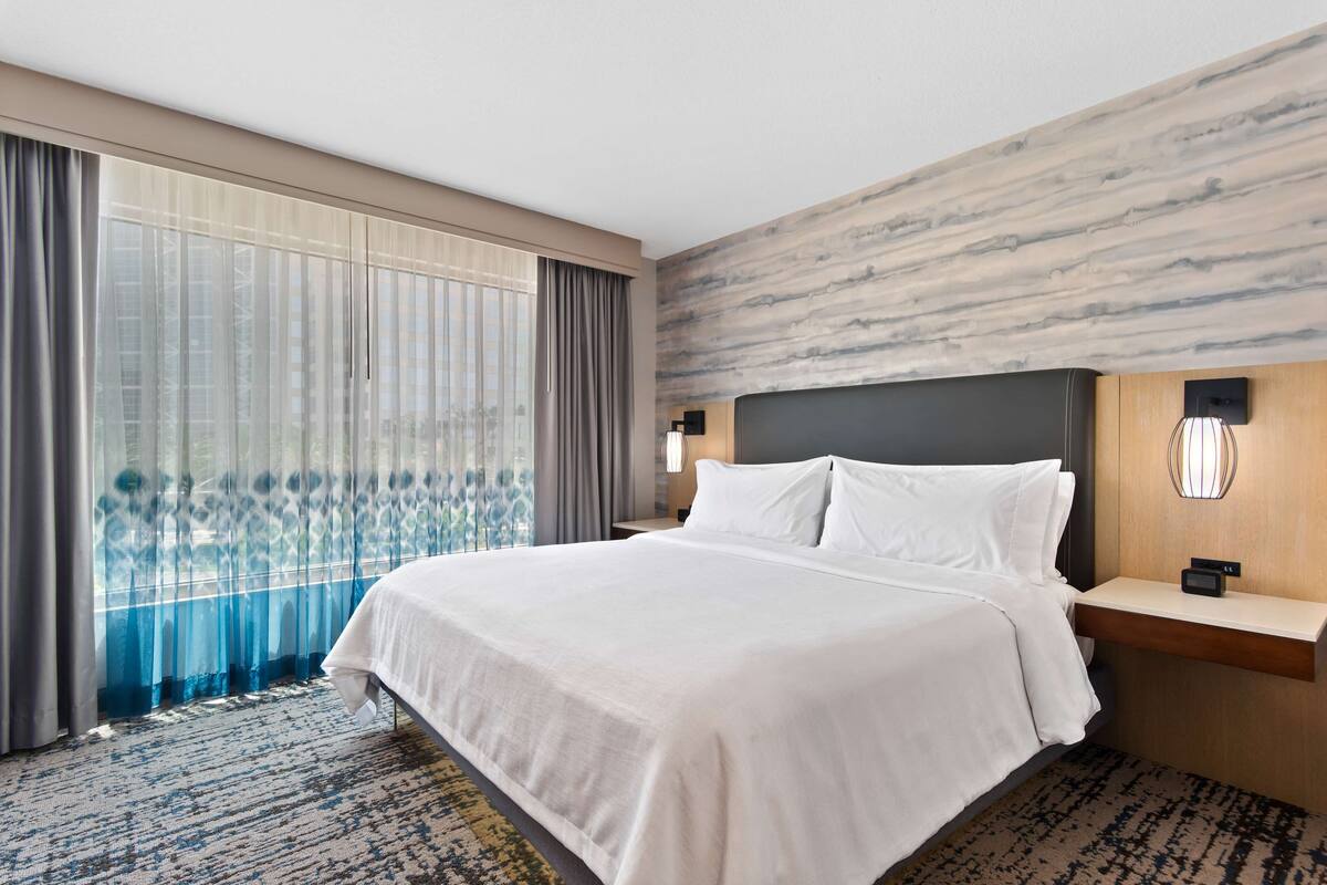A hotel room with a large bed, a nightstand, a wall-mounted lamp, and a view through sheer curtains.