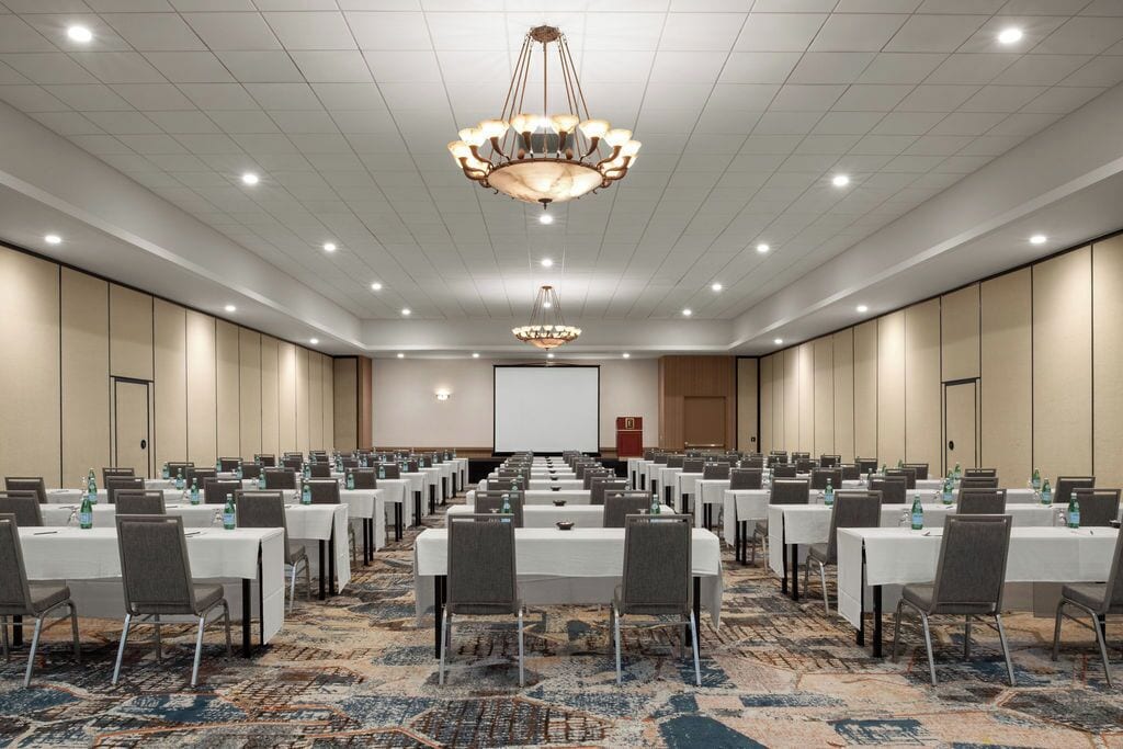 Un salón de conferencias con filas de mesas, sillas y una pantalla grande al frente.
