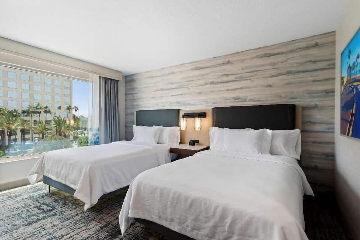 A hotel room with two beds, a large window with a view of palm trees and a building, and a marble-patterned wall.