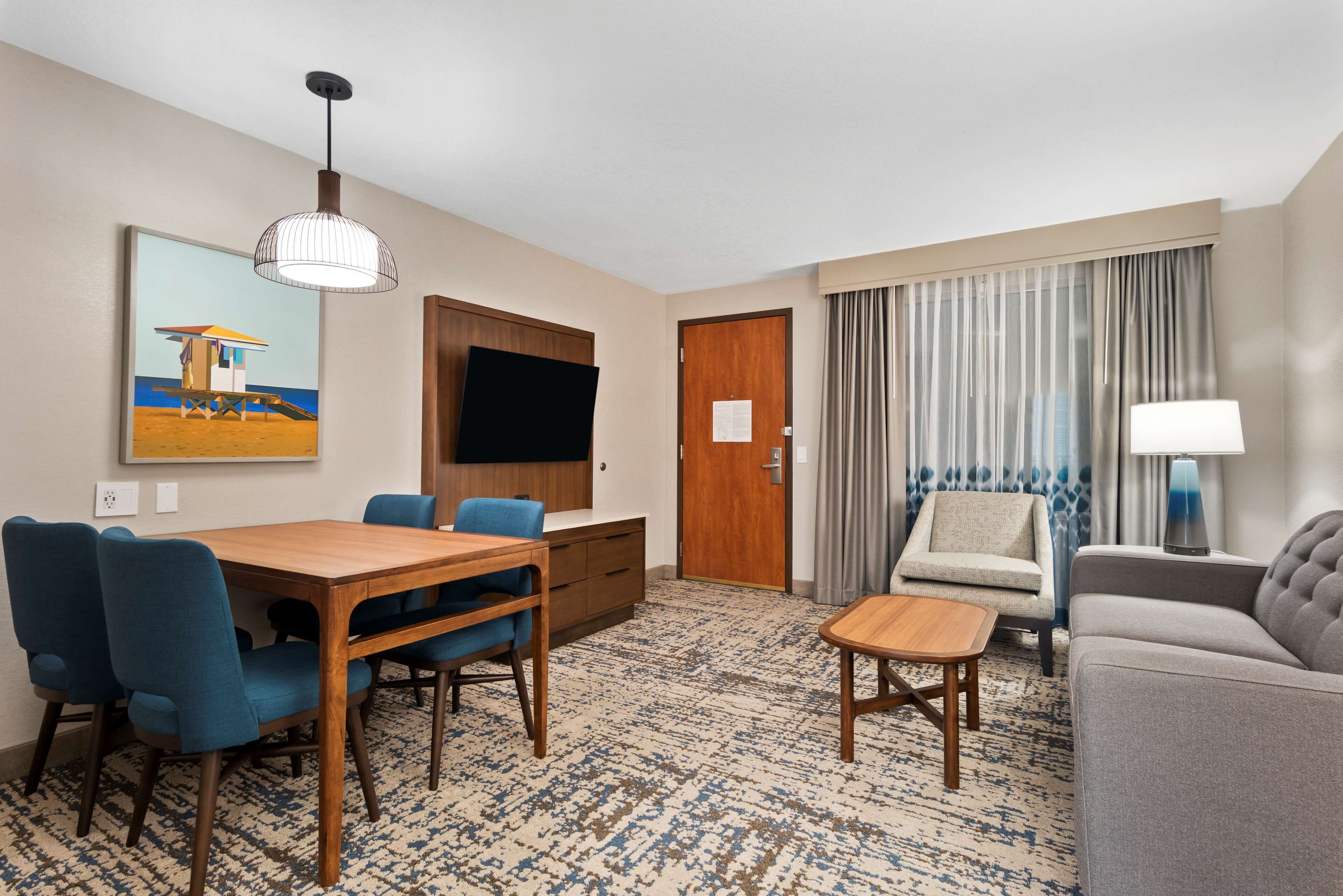 Una habitación de hotel con una mesa de comedor de madera, sillas azules, un sofá gris, una mesita de noche de madera, un televisor montado en la pared y una alfombra estampada.