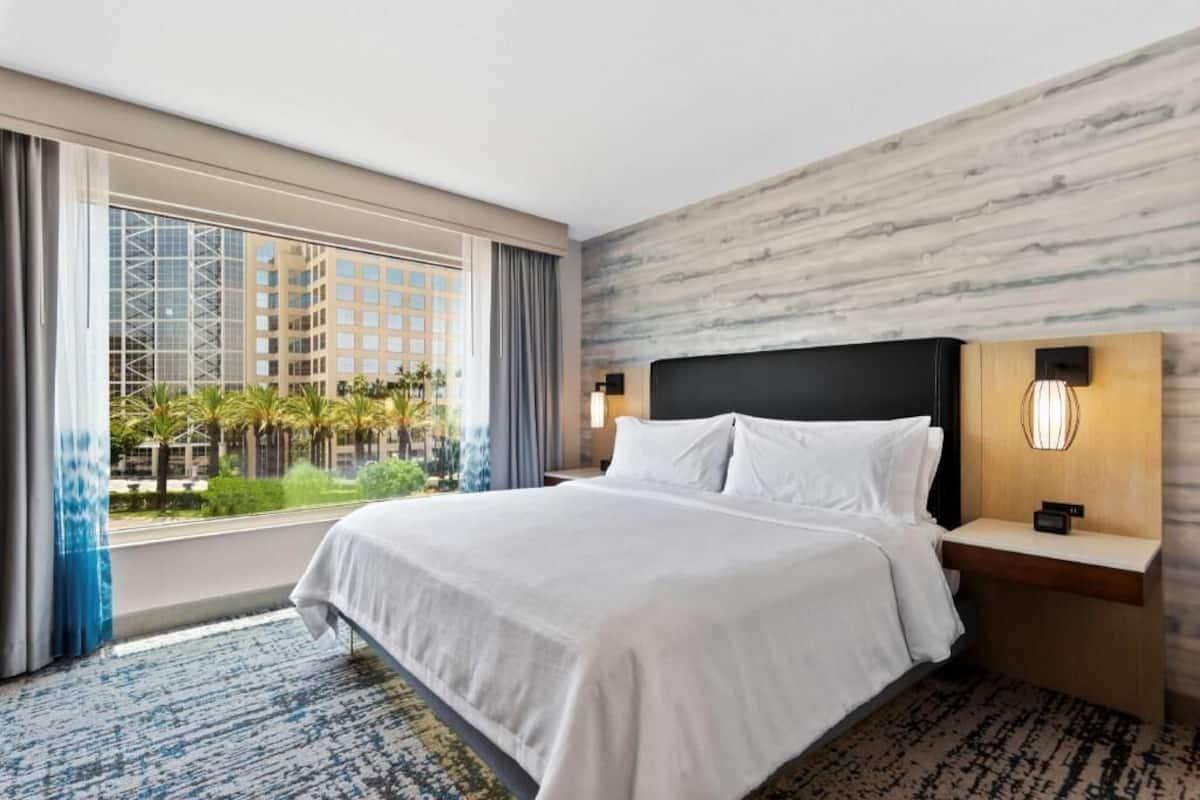 A hotel room with a large bed, a nightstand, a wall-mounted lamp, and a view of a building and palm trees through a window.