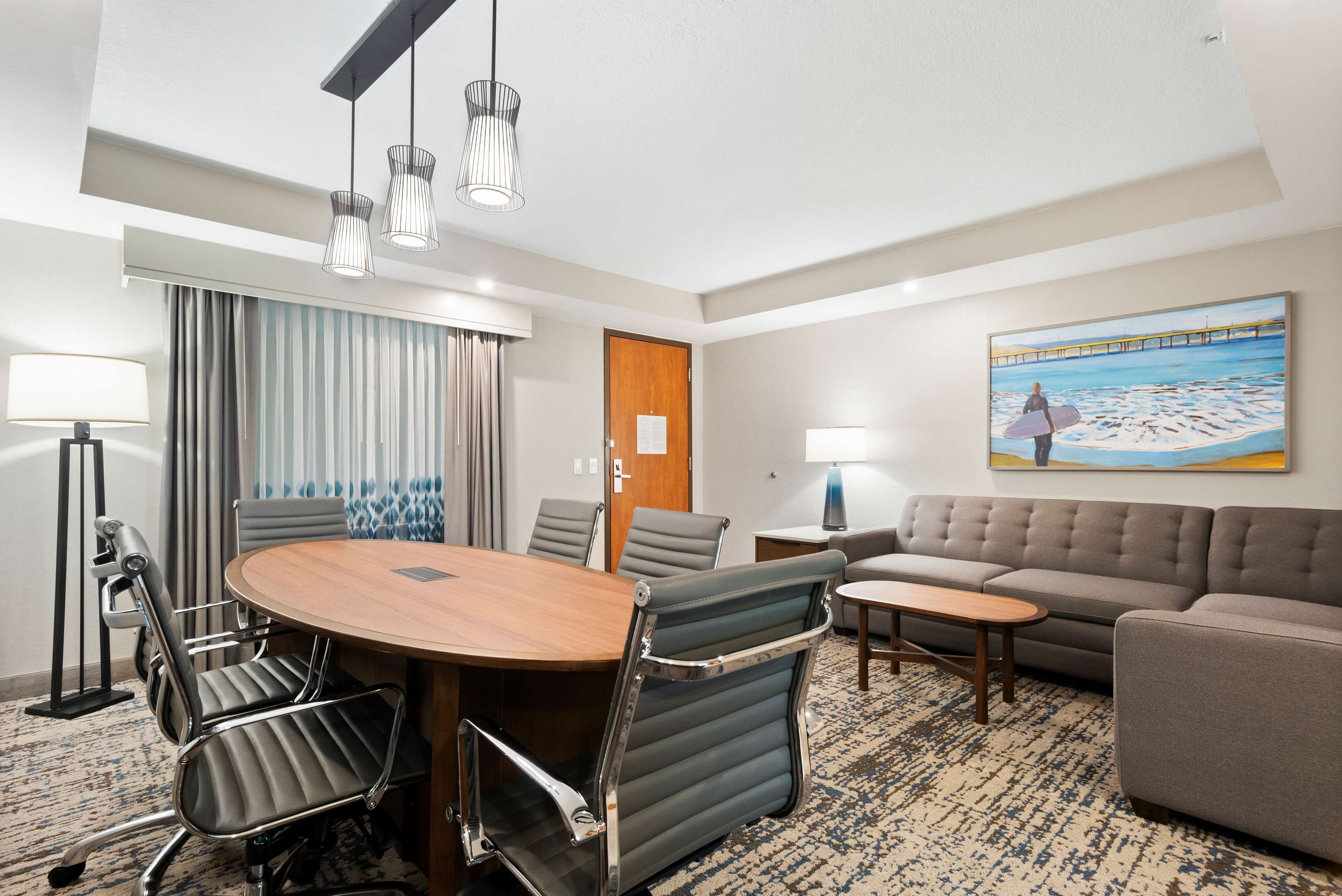 A hotel room with a round wooden table, modern chairs, a sofa, and a painting of a beach scene.
